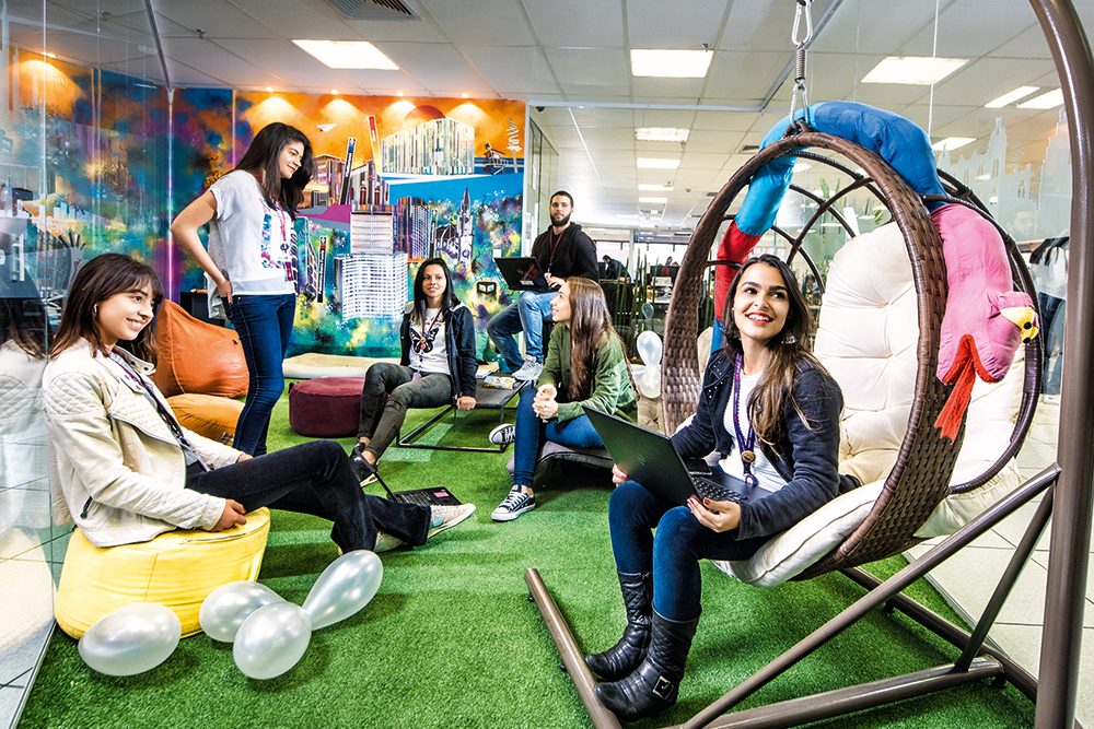 Imagem mostra funcionários em escritório da ClearSale. Eles estão reunidos em um local colorido com grama artificial, balanço e balões