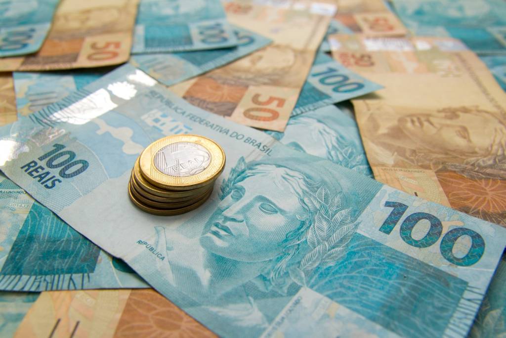 Notas de 100 reais e 50 reais estão espalhadas em uma mesa, onde também há uma pilha de moedas de 1 real