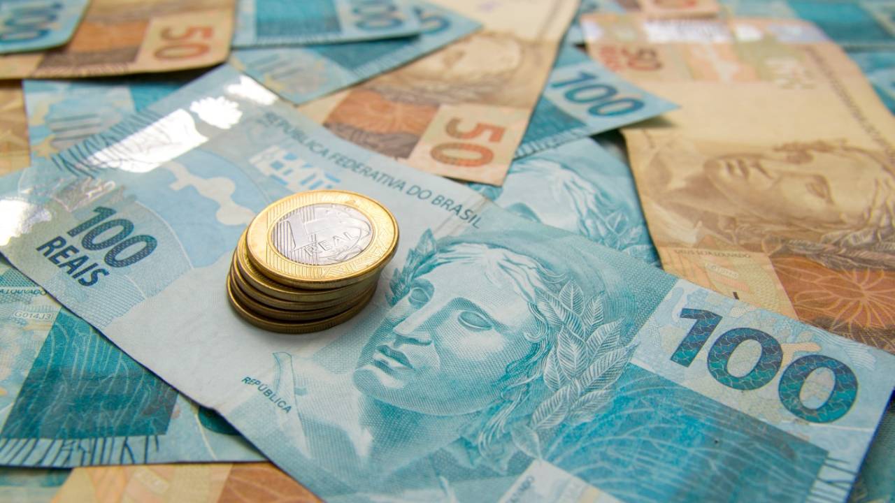 Notas de 100 reais e 50 reais estão espalhadas em uma mesa, onde também há uma pilha de moedas de 1 real