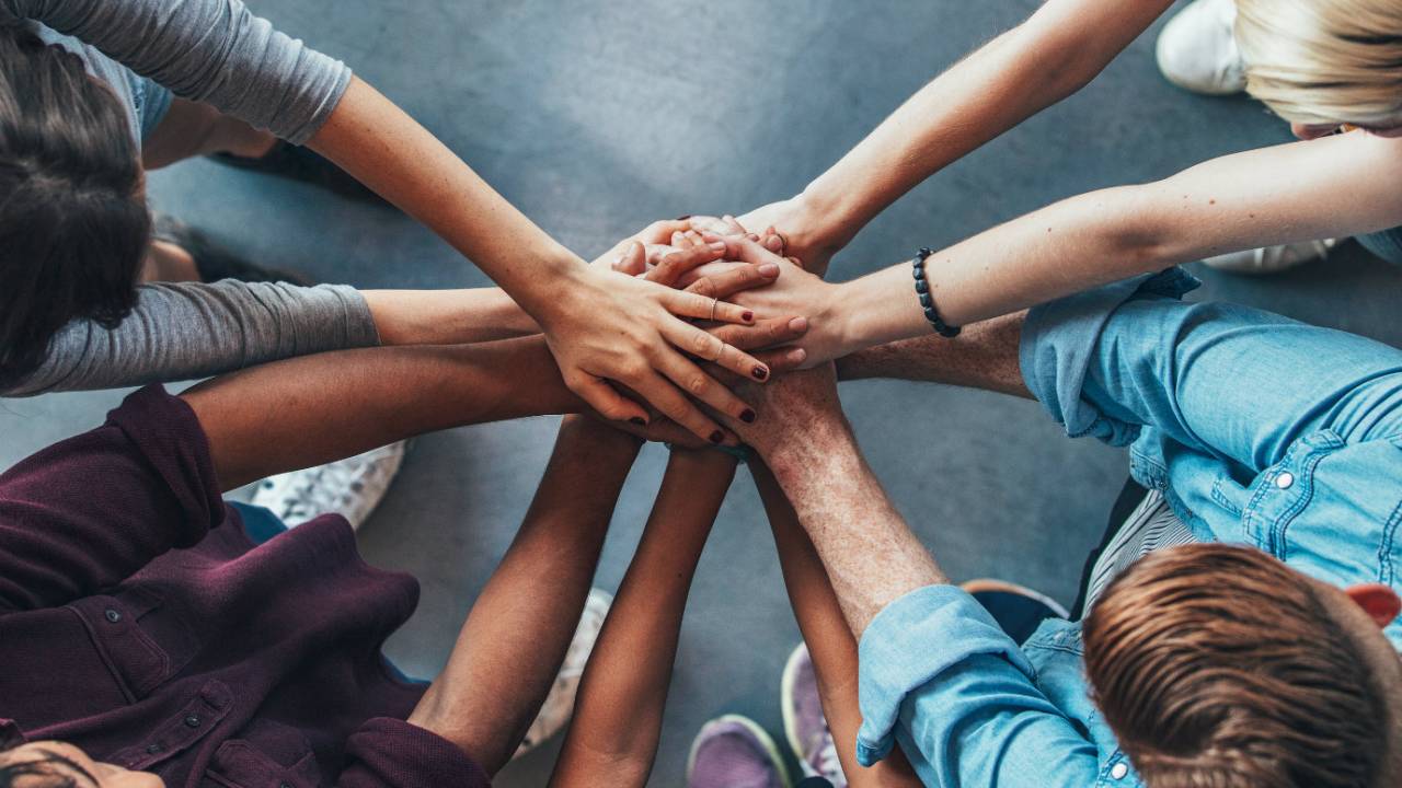 Um grupo de pessoas se reúne em círculo e juntam suas mãos, mostrando colaboração em equipe