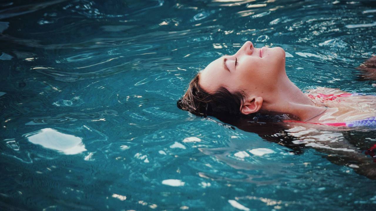 Imagem mostra o rosto de uma mulher branca que está boiando numa piscina.