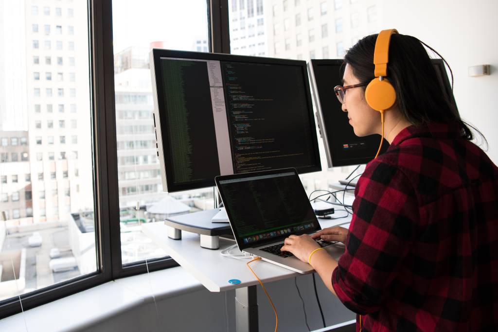 Uma mulher de aproximadamente 25 anos aparece trabalhando usando o computador com mais dois monitores conectados a ele, enquanto ela ouve música em fones de ouvido amarelos.