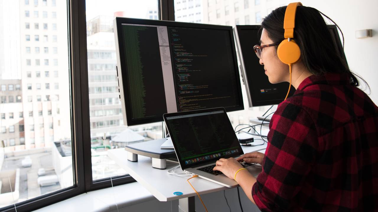 Uma mulher de aproximadamente 25 anos aparece trabalhando usando o computador com mais dois monitores conectados a ele, enquanto ela ouve música em fones de ouvido amarelos.
