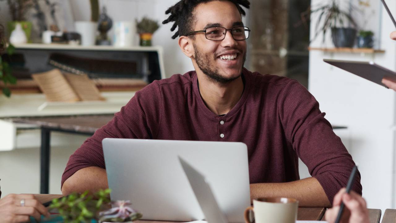 Um homem negro de aproximadamente 25 anos aparece sorrindo em frente a um computador.