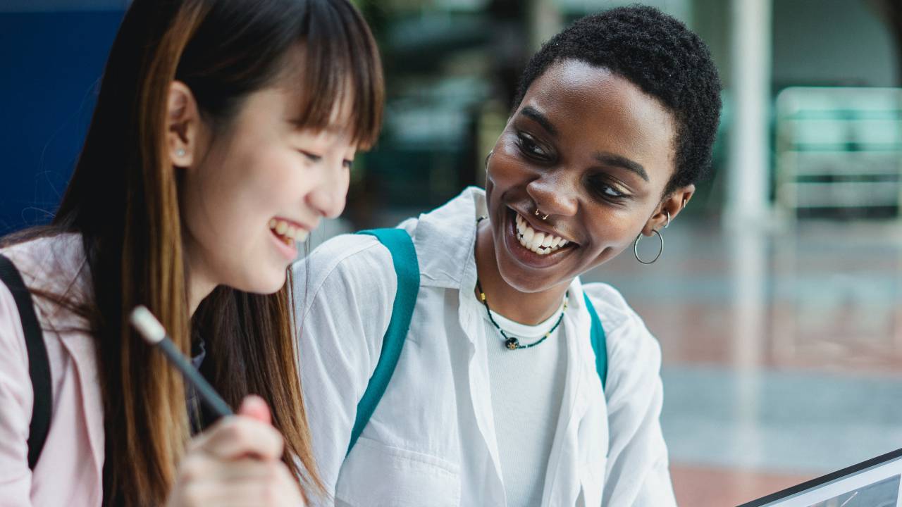 Uma jovem negra de aproximadamente 20 anos olha para sua colega asiática, também de aproximadamente 20 anos. Ambas sorriem.