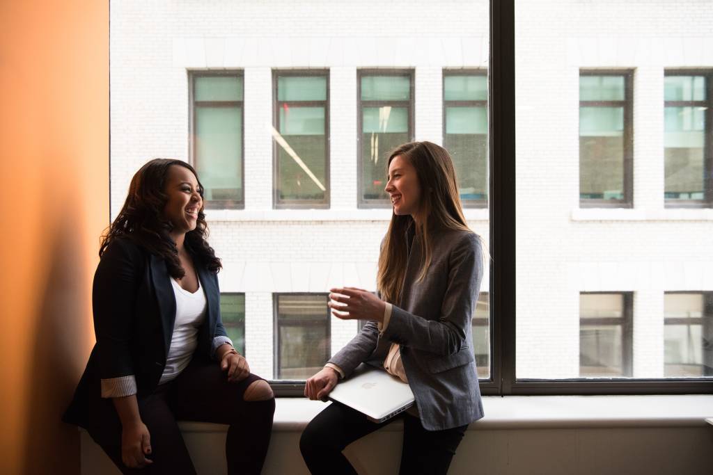 Duas mulheres aparecem sentadas próximas a uma janela, dentro de um escritório. Elas sorriem enquanto conversam.