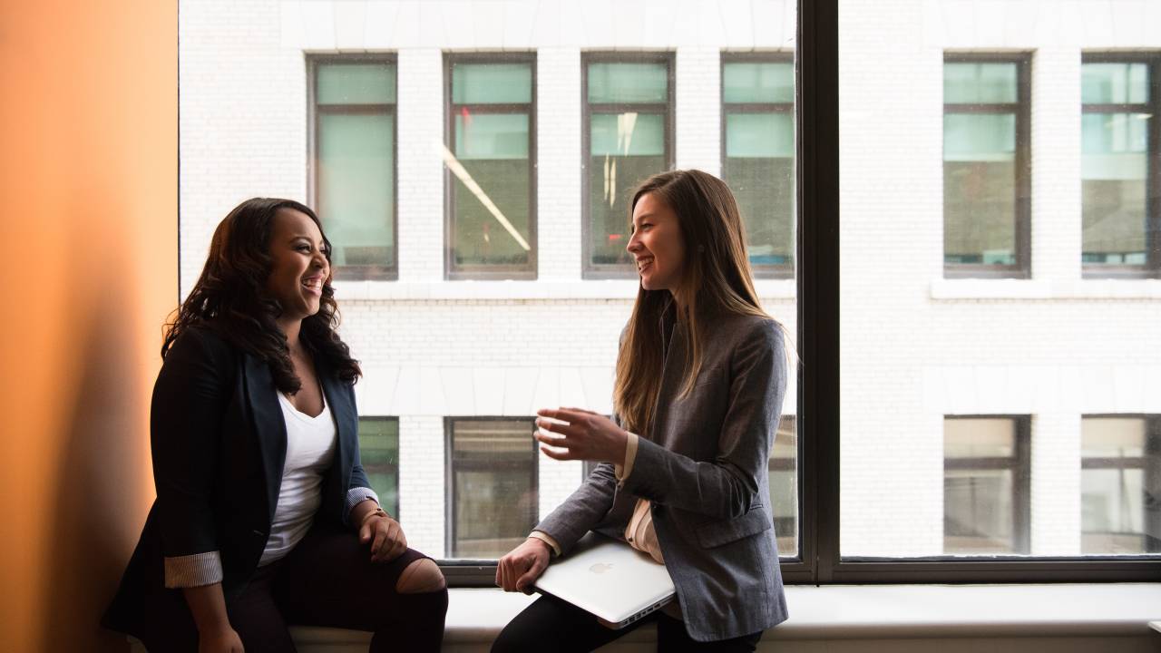 Duas mulheres aparecem sentadas próximas a uma janela, dentro de um escritório. Elas sorriem enquanto conversam.