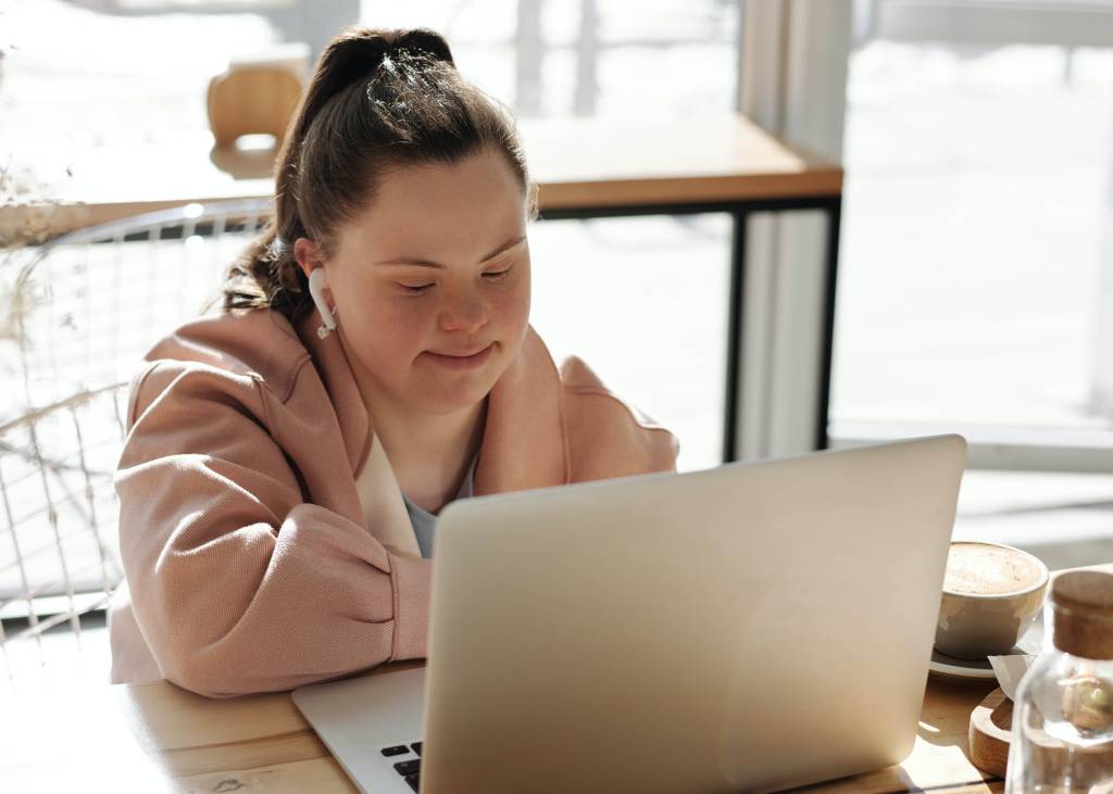 Uma jovem branca, de aproximadamente 25 anos, com síndrome de down, aparece sentada, trabalhando em frente a um computador.