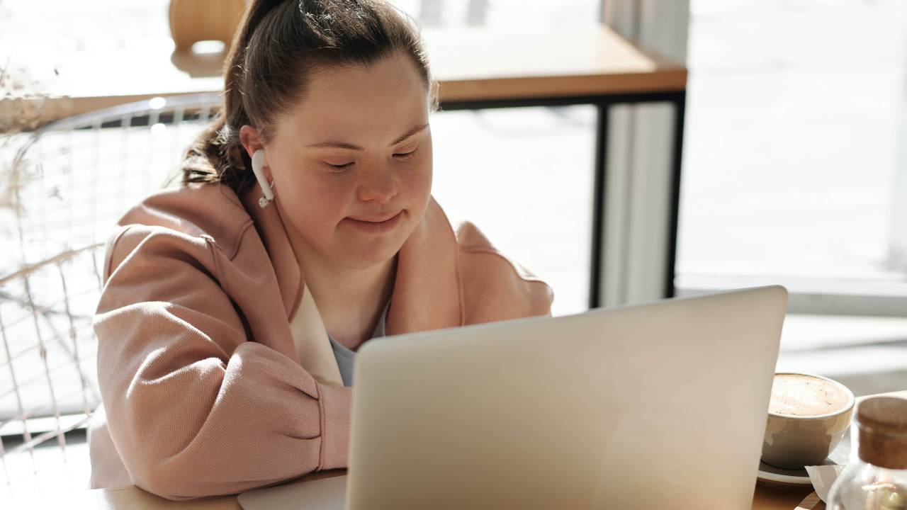 Uma jovem branca, de aproximadamente 25 anos, com síndrome de down, aparece sentada, trabalhando em frente a um computador.