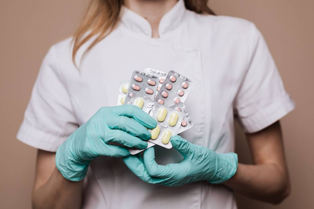 Imagem de uma com roupas hospitalares, usando luvas e segurando embalagens de remédio.