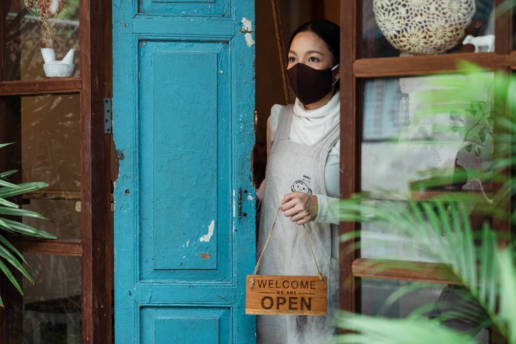 Mulher oriental com máscara colocando a placa "open" na porta de um negócio.