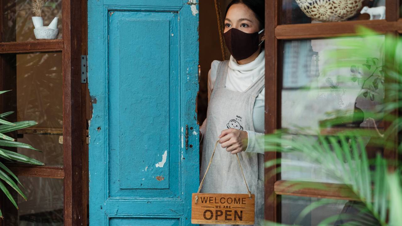 Mulher oriental com máscara colocando a placa "open" na porta de um negócio.