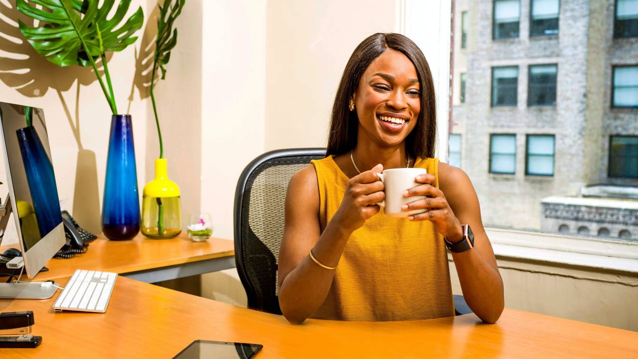 Mulher negra tomando café e sorrindo num escritório