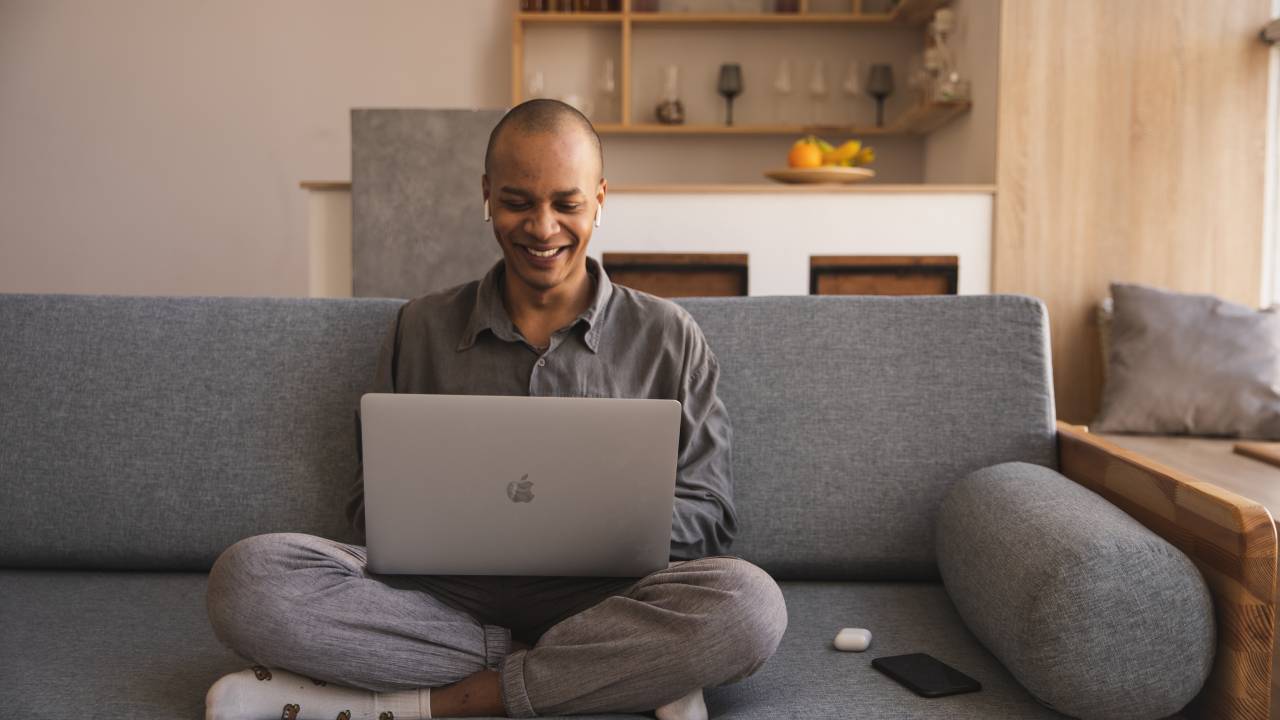 Um homem aparece sentado no sofá sorrindo enquanto usa o computador.