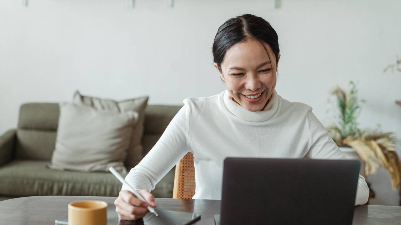 Uma mulher aparece sentada e trabalhando no computador, enquanto sorri.
