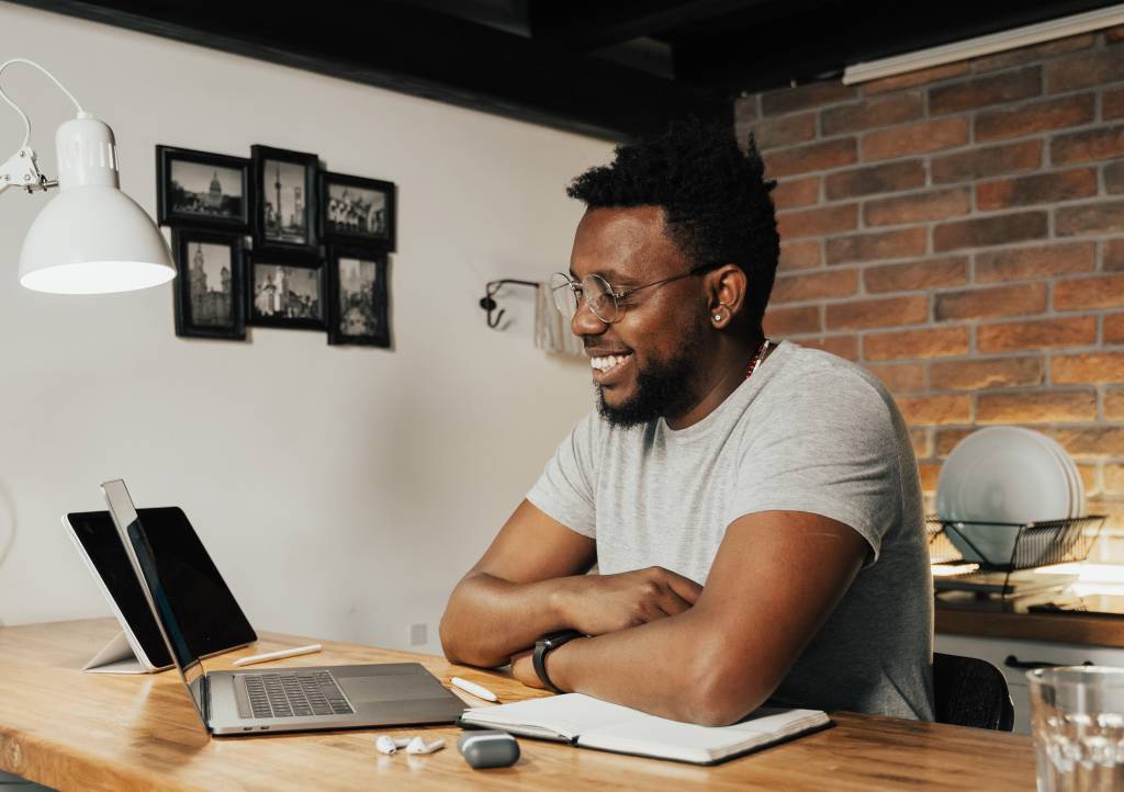 Um homem negro de aproxidamente 30 anos aparece em sua mesa de trabalho sentado em frente a um computador. Na imagem, ele sorri.