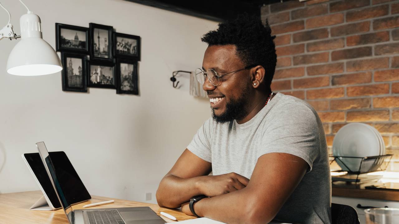 Um homem negro de aproxidamente 30 anos aparece em sua mesa de trabalho sentado em frente a um computador. Na imagem, ele sorri.