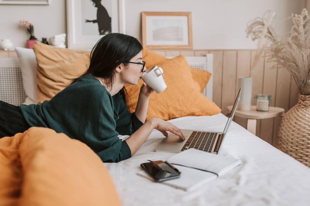 Mulher de óculos deitada na cama tomando café e estudando no notebook.