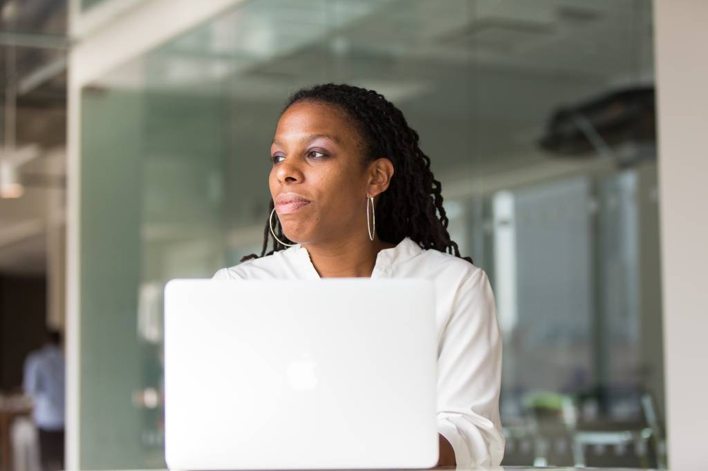 Imagem mostra uma mulher negra sentada atrás de um notebook branco e olhando para o horizonte