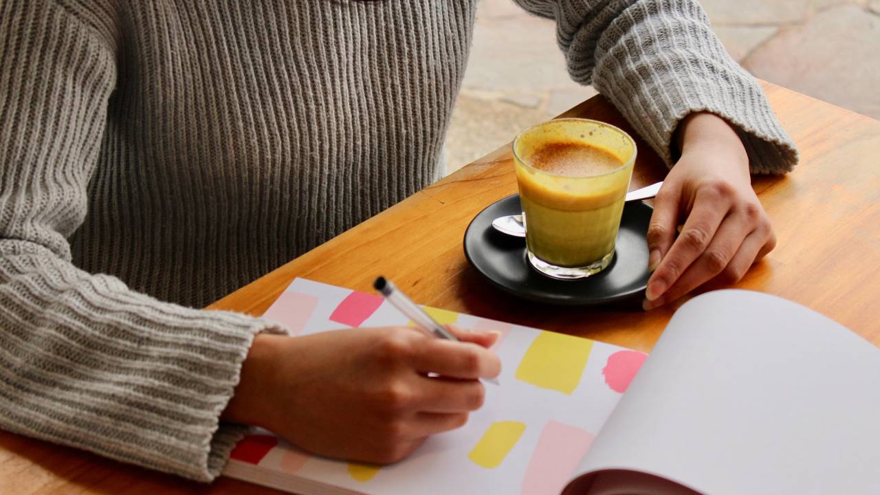 Imagem mostra as mãos de uma mulher escrevendo em um bloco de páginas brancas com quadrados coloridos (rosa e amarelo). Na imagem também existe um copo com capuccino.