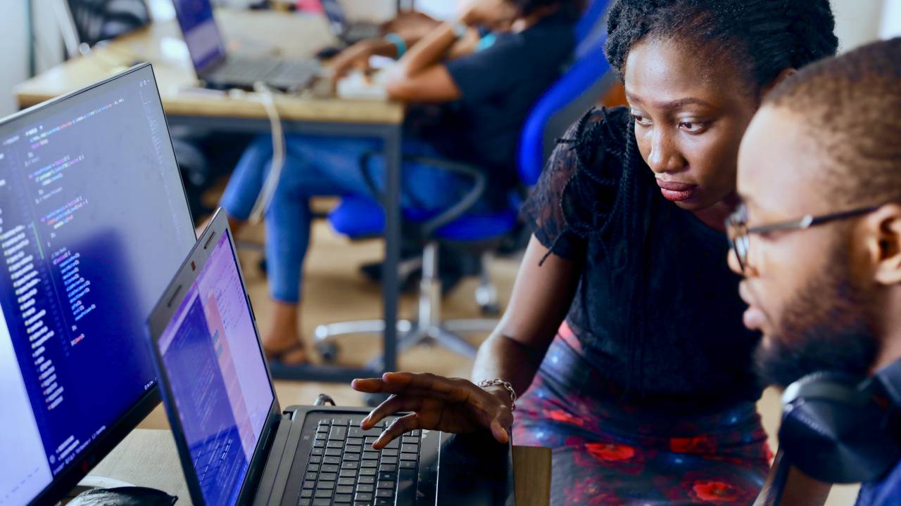 A imagem mostra um homem negro sentado em frente a um computador e, ao seu lado, uma mulher negra em pé está olhando para a tela e analisando o trabalho