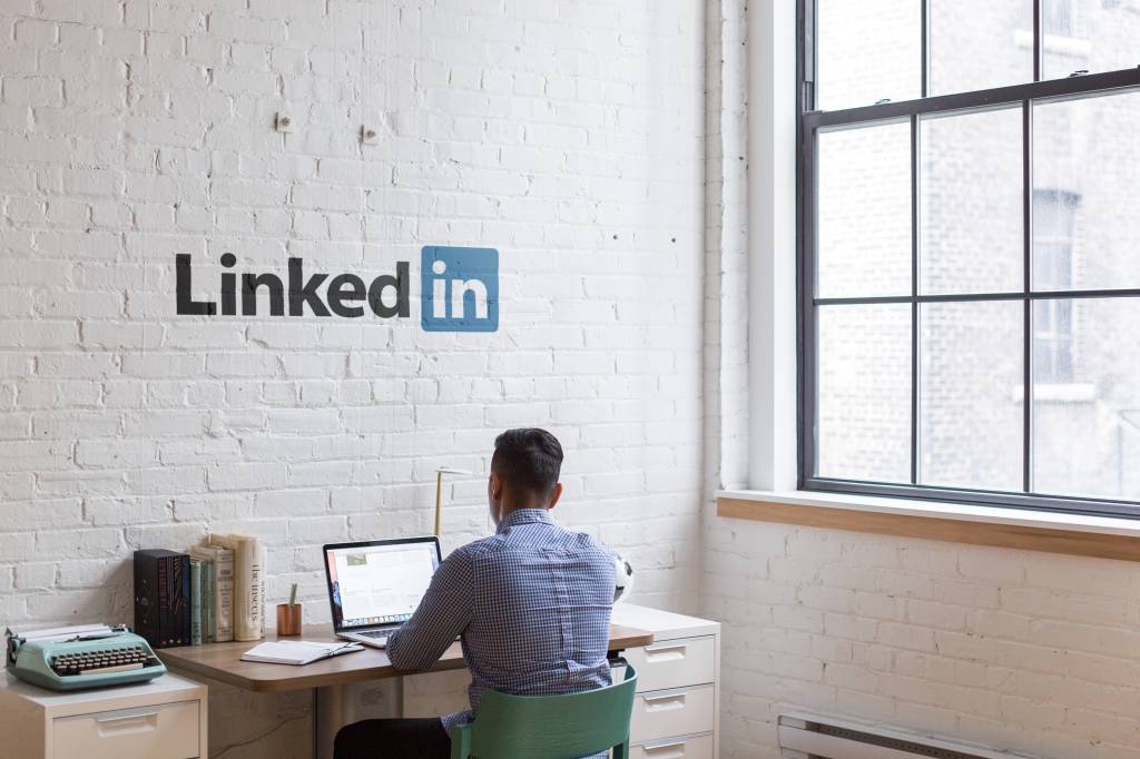 Foto mostra um homem de cabelos pretos e camisa azul de costas trabalhando em um notebook. Na parede acima dele está o símbolo do LinkedIn.