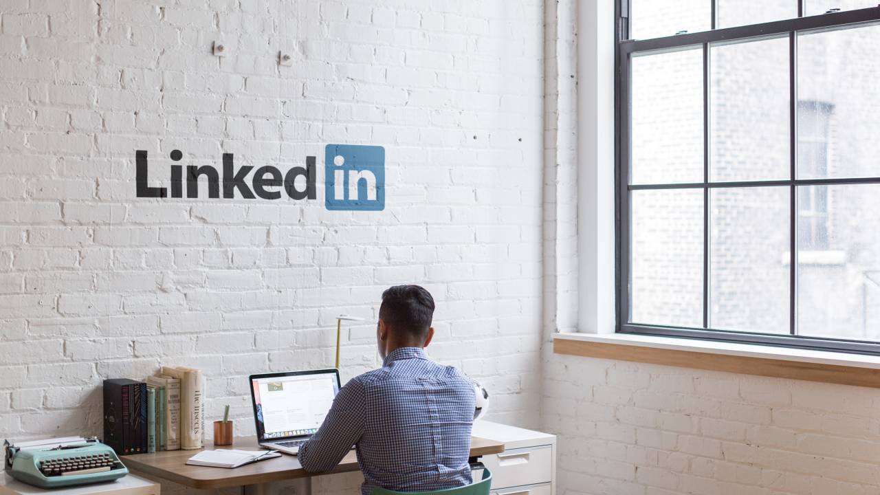 Foto mostra um homem de cabelos pretos e camisa azul de costas trabalhando em um notebook. Na parede acima dele está o símbolo do LinkedIn.
