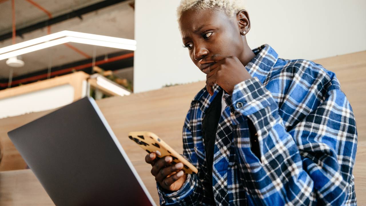 A foto mostra uma mulher negra de cabelos curtos e loiros olhando apreensiva para o computador com um celular nas mãos.