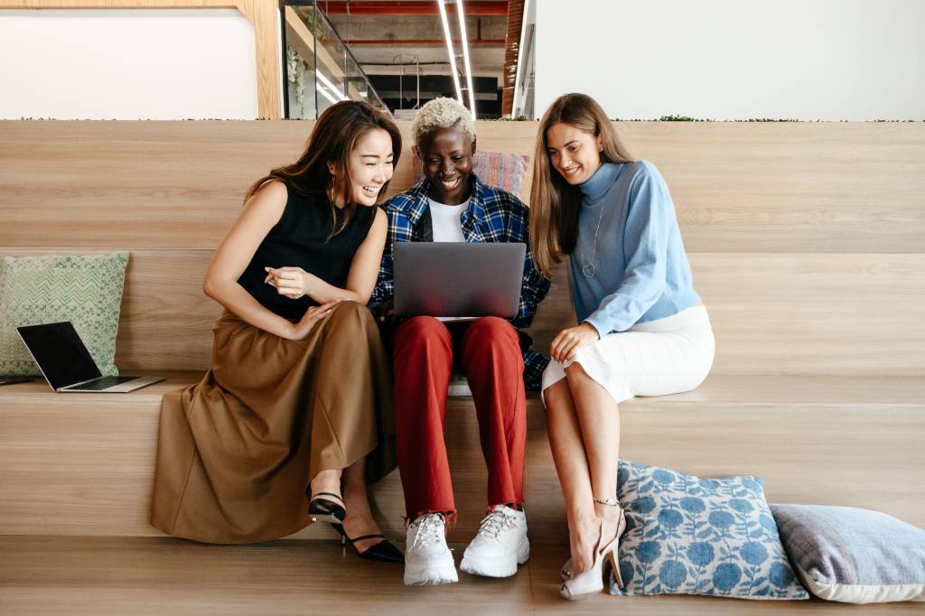 Três mulheres (uma oriental, uma negra e uma loira) trabalhando juntas sentadas juntas olhando o notebook de uma delas e sorrindo.