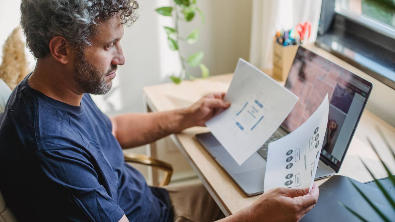 Imagem mostra home grisalho sentado em frente a um notebook olhando para folhas de papel