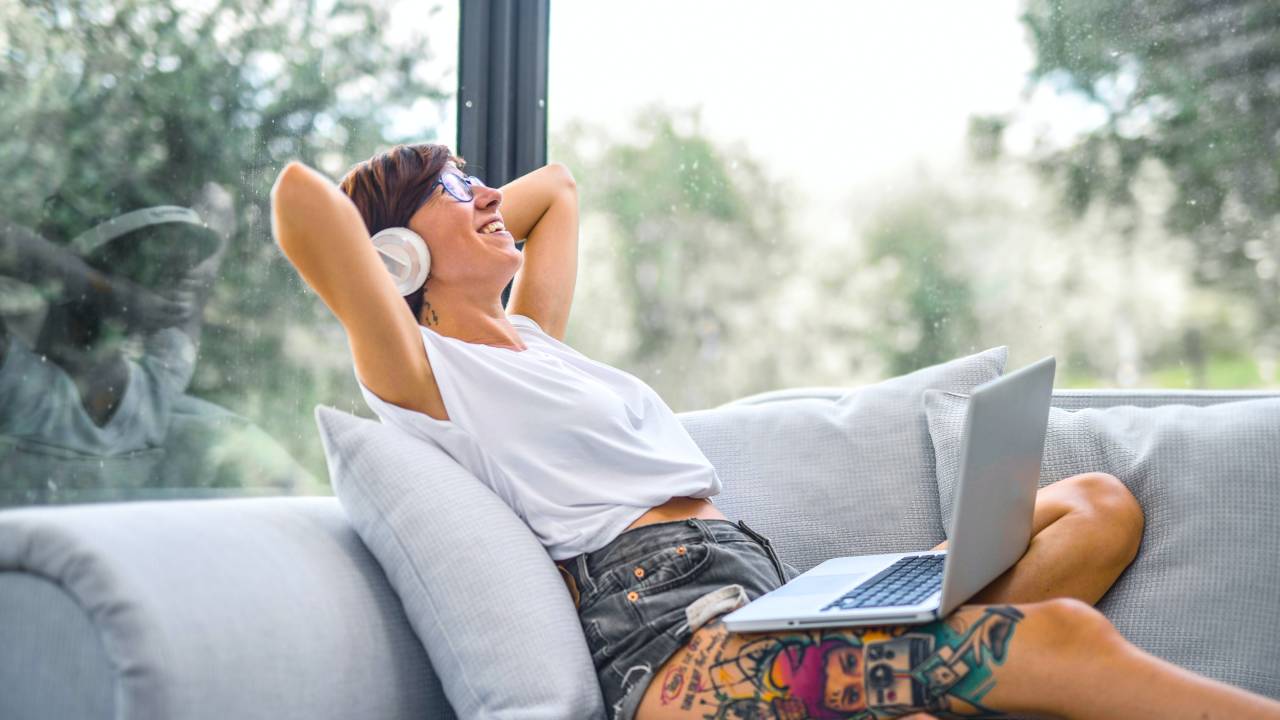 Mulher de cabelos curtos e tatuagem sentada em um sofá com os braços atrás da cabeça, notebook no colo e fone de ouvido, sorrindo.