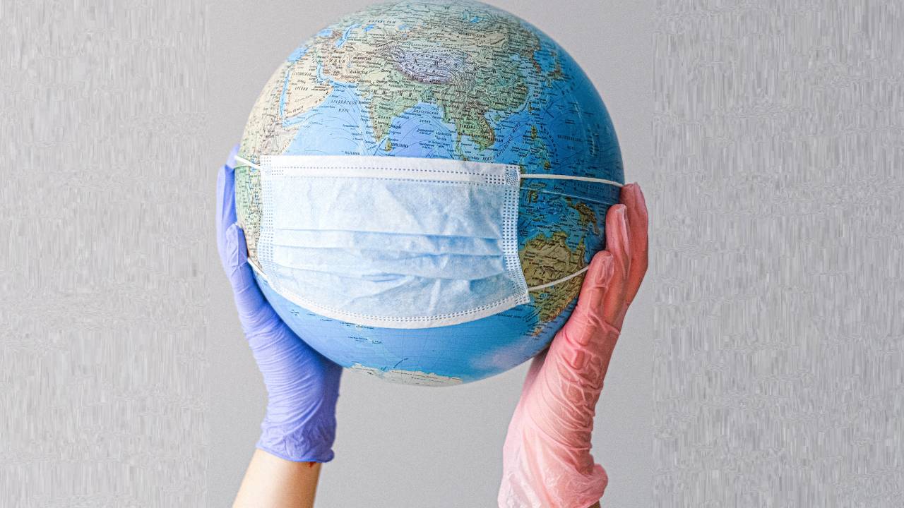 Duas mãos com luvas cirúrgicas aparecem segurando um globo terrestre com uma máscara também cirúrgica.