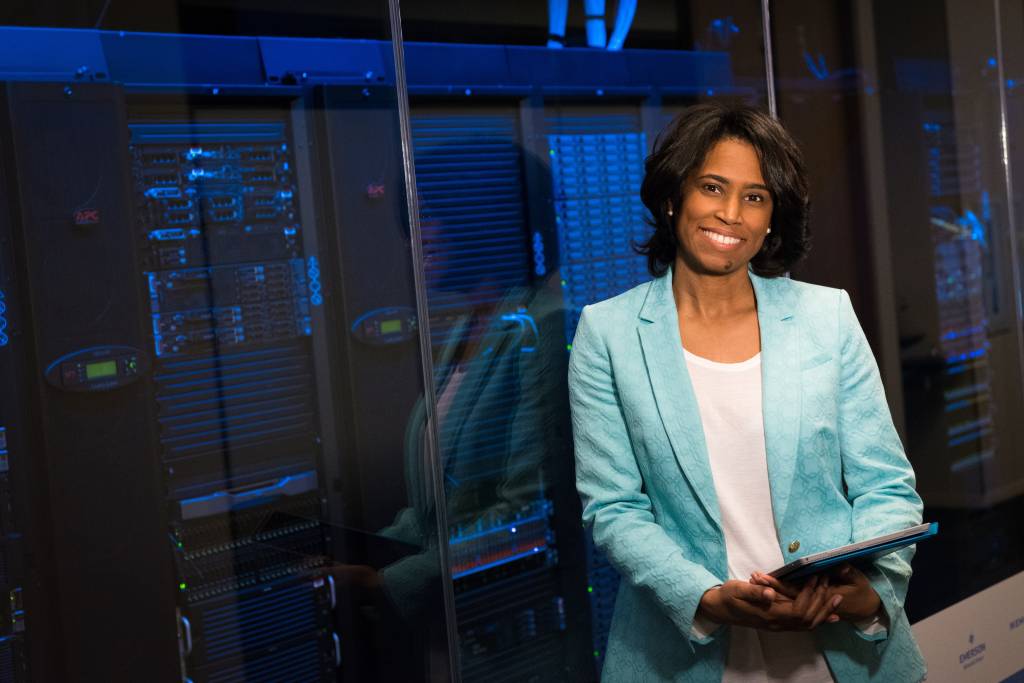 Foto mostra uma mulher negra sorrindo, segurando um tablet, em frente a um data-center