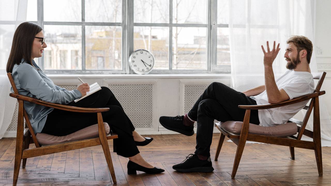 Foto mostra uma mulher de camisa azul que é psicóloga atendendo um homem de camisa branca. Os dois estão sentados frente a frente.