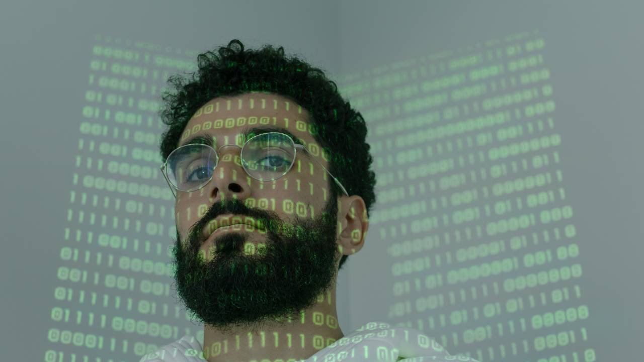 Um homem de aproximadamente 30 anos, usando óculos de grau aparece com uma projeção de algoritmos em seu rosto e na parede em seu fundo.
