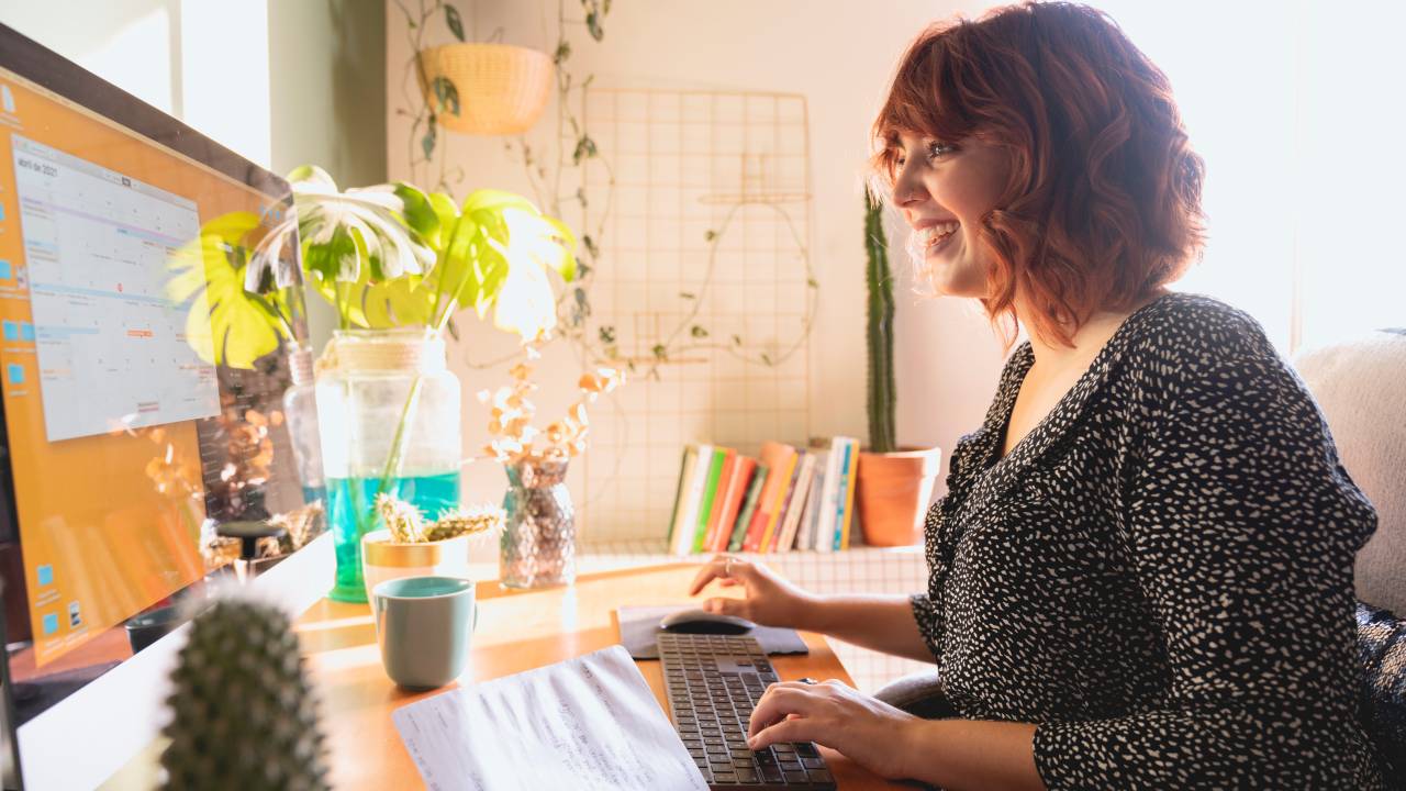 Imagem mostra uma mulher de cabelos ruivos trabalhando em home office iluminado e cercado por plantas e sorrindo