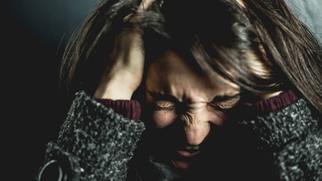 Imagem mostra mulher de cabelos escuros com os olhos fechados, mãos na cabeça e expressão de raiva.