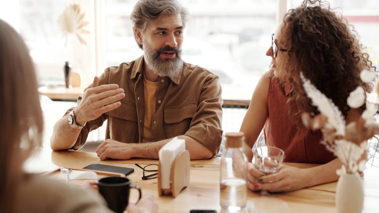 Um homem de barba e uma mulher de cabelos cacheados conversam em uma mesa de reunião.