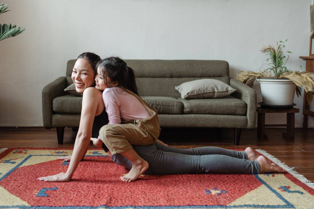 Uma mulher aparece realizando um movimento de ioga no tapete de sua sala , enquanto recebe um abraço de sua filha criança.