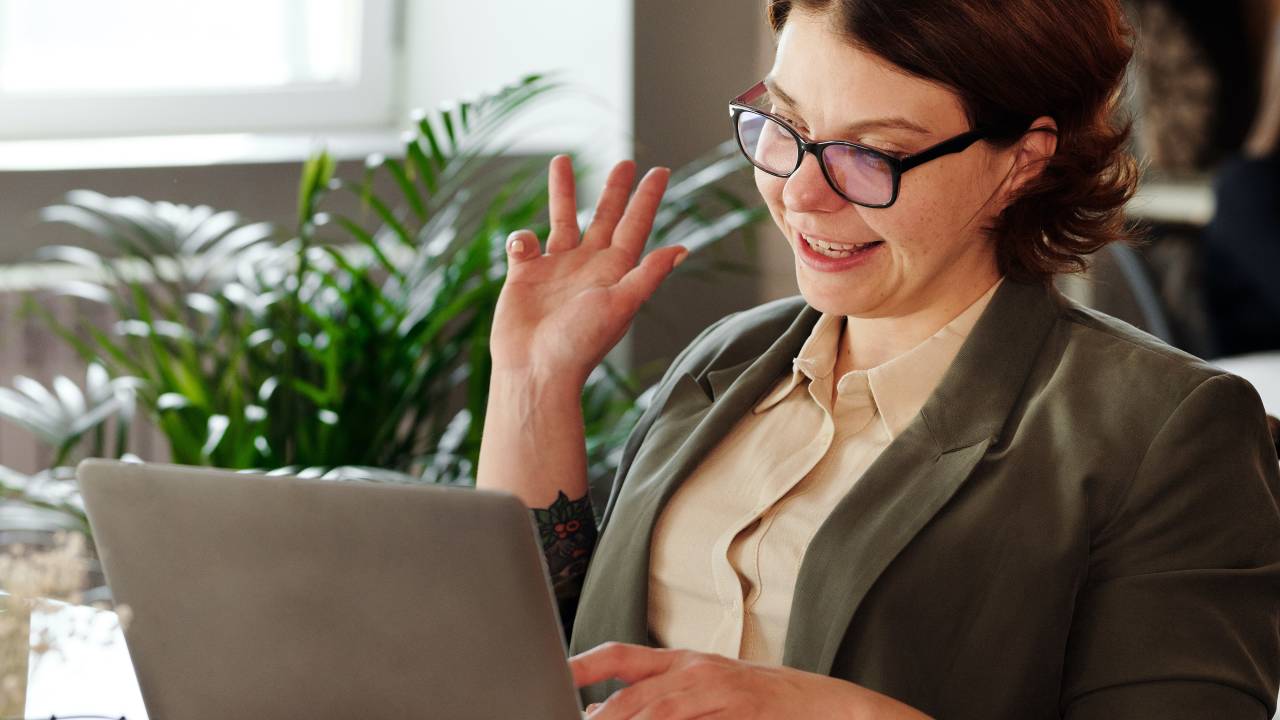 Uma mulher aparece sorrindo em frente a um computador, aparentando estar em uma vídeo-chamada.