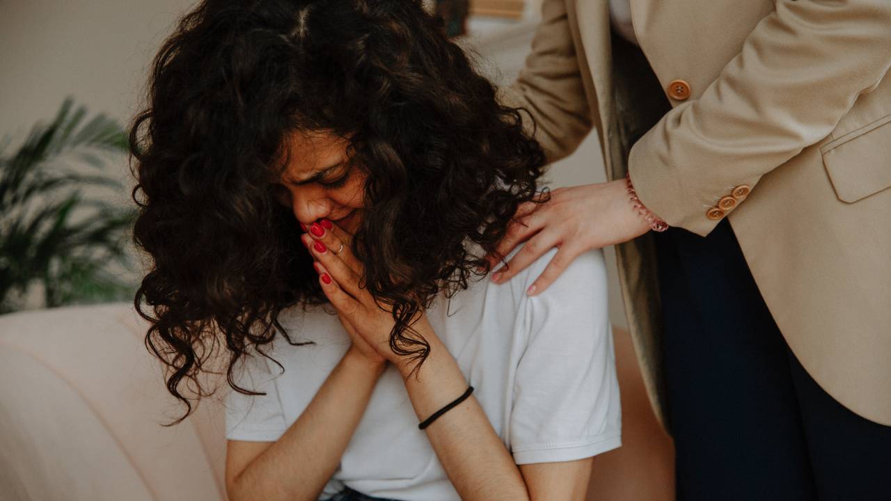 Imagem mostra jovem de cabelos cacheados chorando com o rosto baixo parte de um corpo de mulher que apoia as maos nos ombros da jovem.