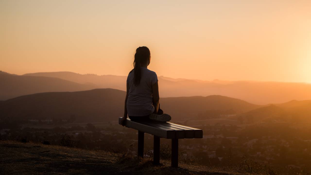 Imagem mostra uma mulher de costas sentada em um banco em frente a montanhas.