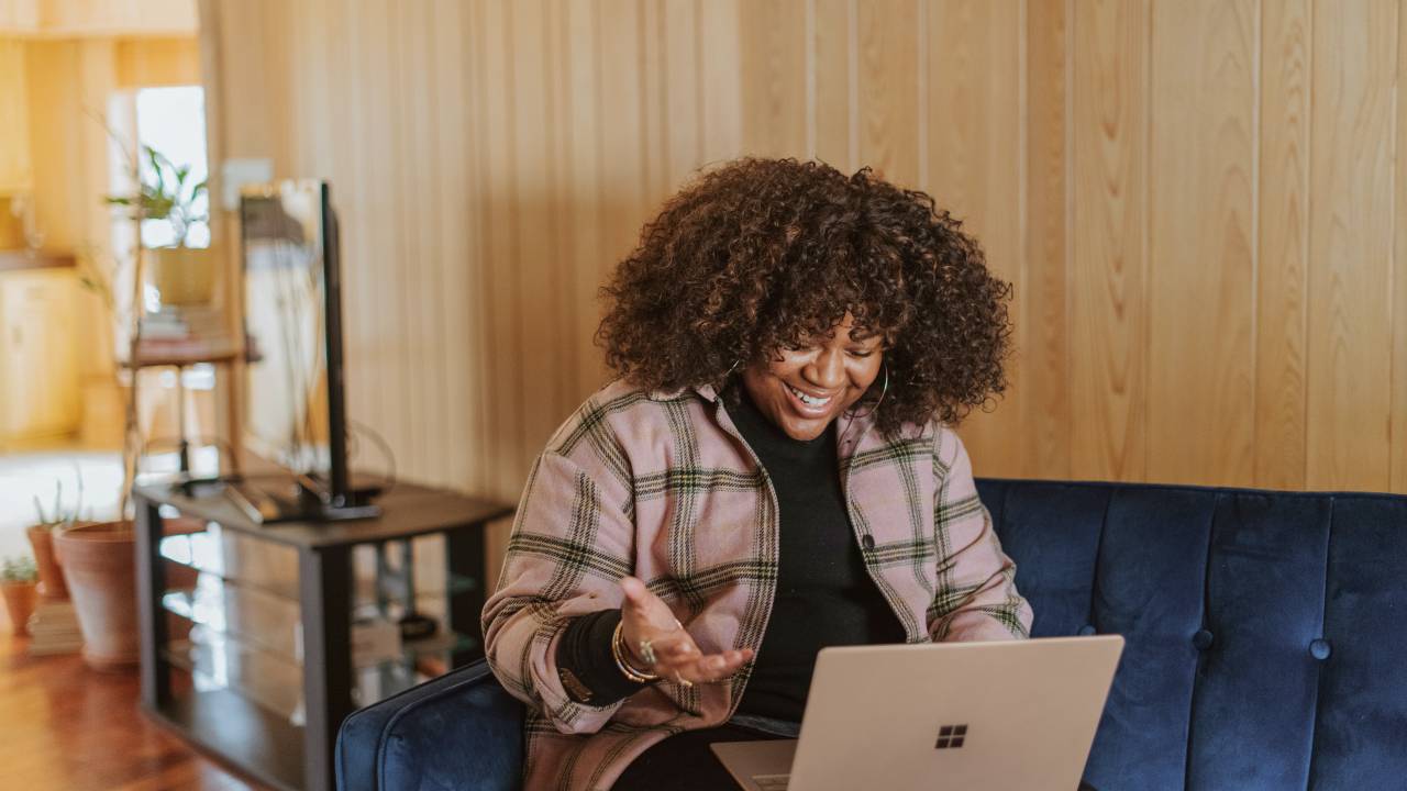 Imagem mostra mulher negra sentada em um sofá com notebook no colo em uma videoconferência