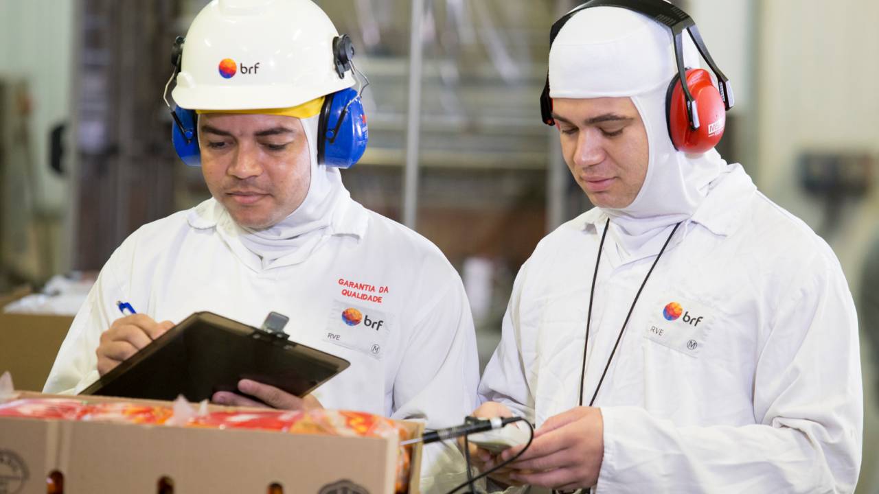 Imagem mostra dois funcionários da BRF com uniforme brancos e capacetes na linha de produção