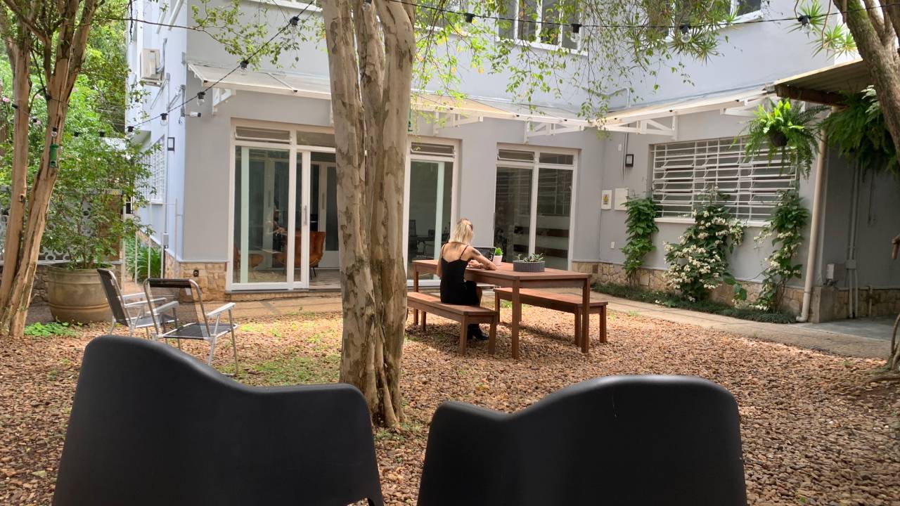 Imagem mostra o jardim de uma casa com árvores. Uma mulher loira está de costas sentada numa mesa no meio do jardim.