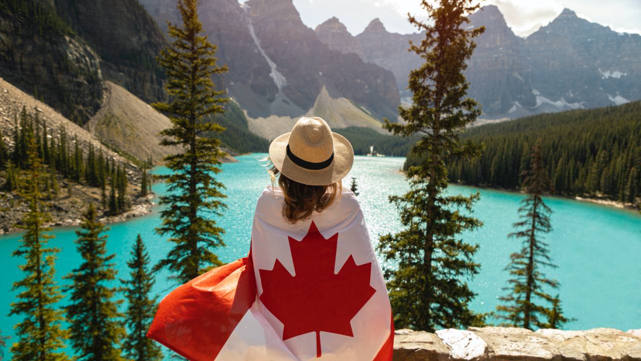Imagem mostra mulher de costas enrolada em um bandeira do Canadá olhando para um lago.