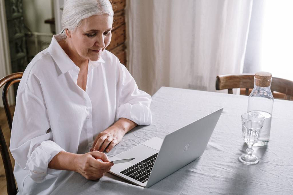 Imagem mostra uma mulher de cabelos grisalhos e camisa branca em frente a um notebook.
