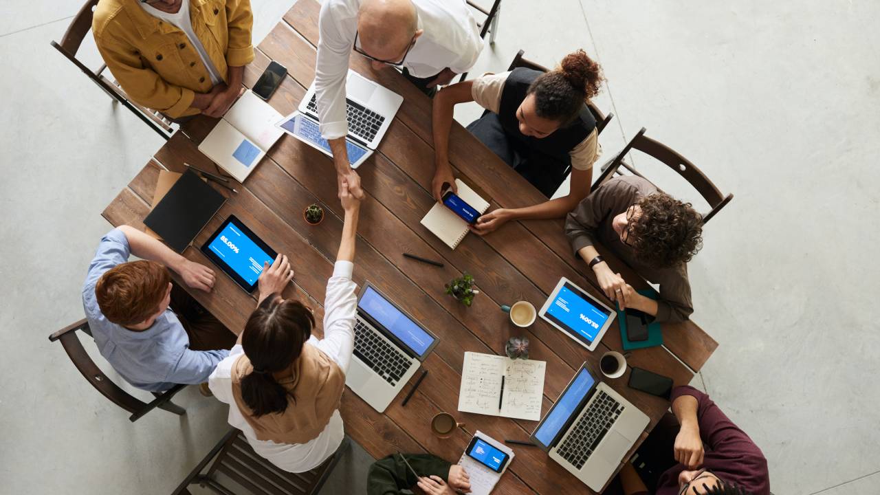 Imagem mostra uma mesa de reunião vista de cima com pessoas trabalhando. Duas delas apertam as mãos.