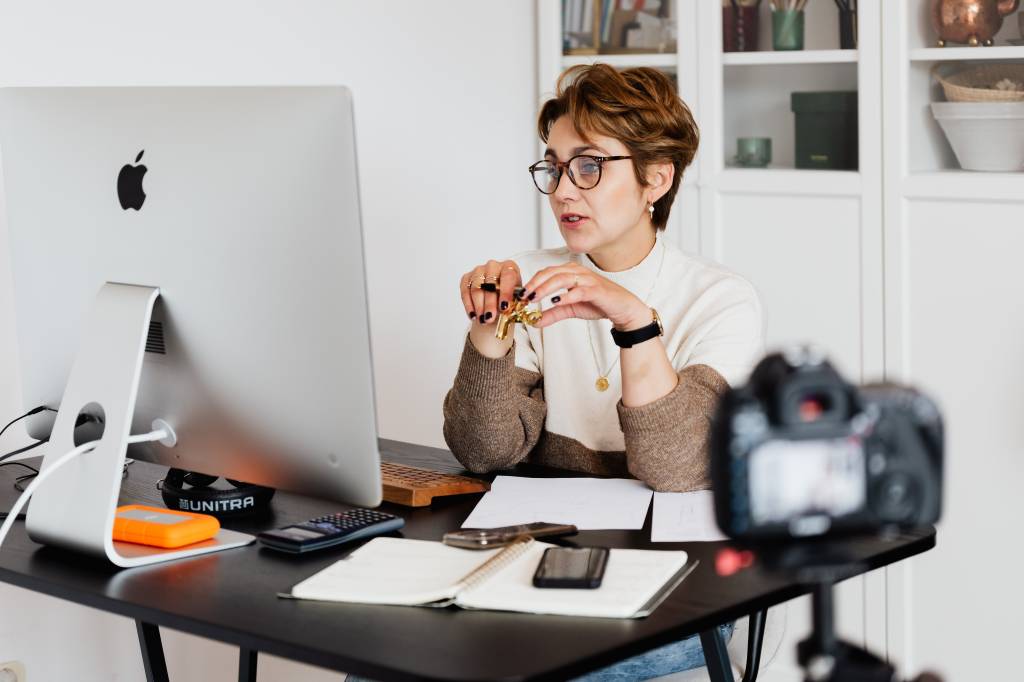 Imagem mostra uma mulher branca, de cabelos curtos, sentada em frente a um monitor, conversando com alguém remotamente.