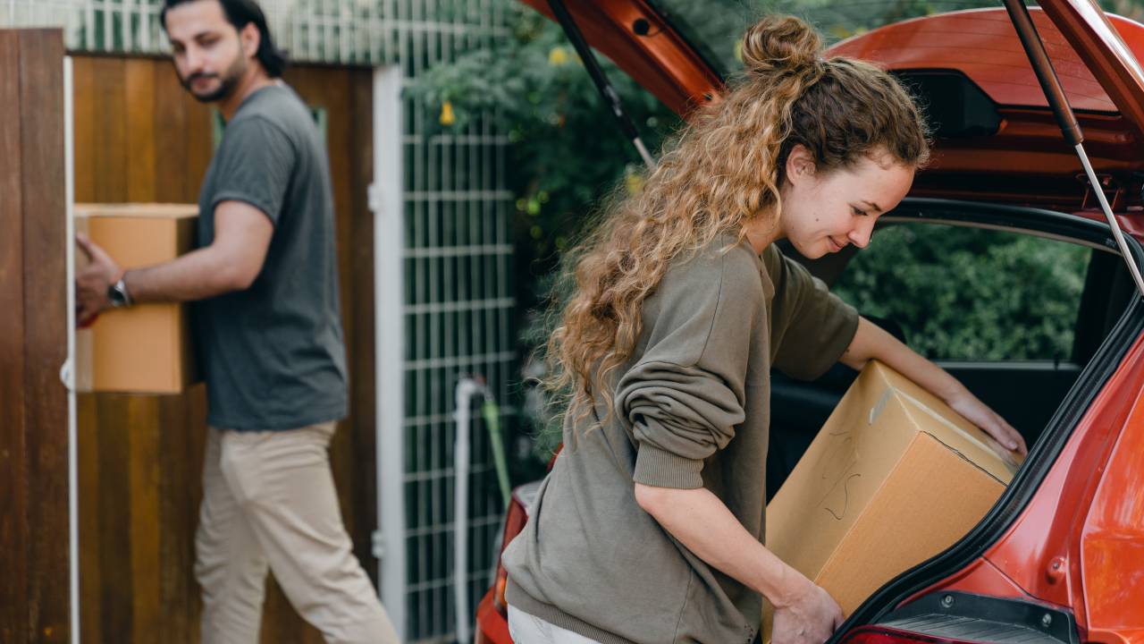 Imagem mostra um homem e uma mulher brancos carregando caixas de mudança. Ela está tirando uma caixa de dentro do porta-malas de um carro vermelho.